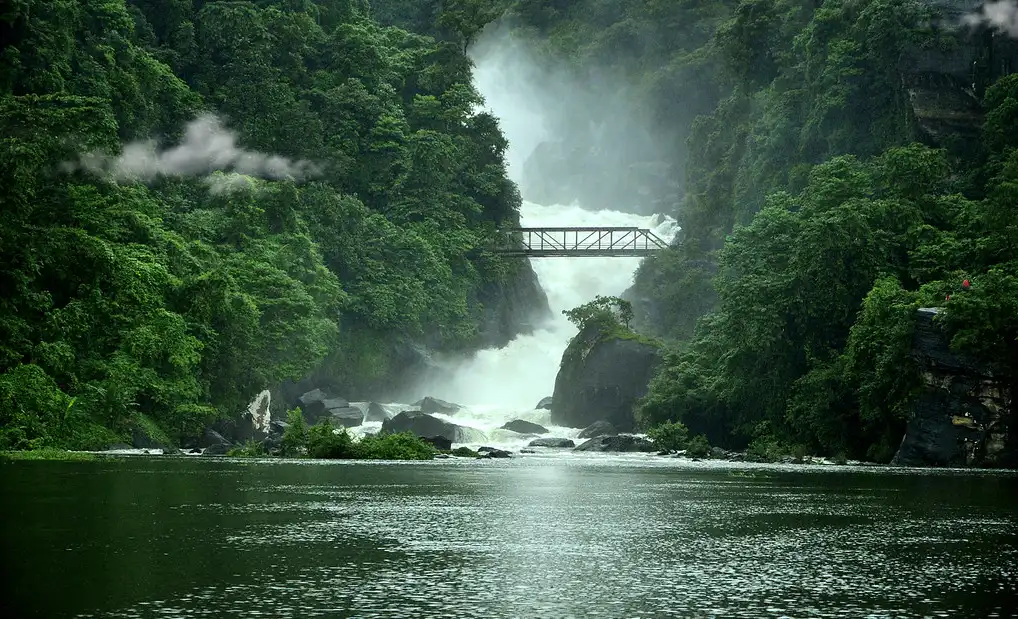 A lush, green forest surrounds a powerful waterfall cascading under a metal bridge. Mist rises, creating an ethereal, tranquil atmosphere over the calm river.