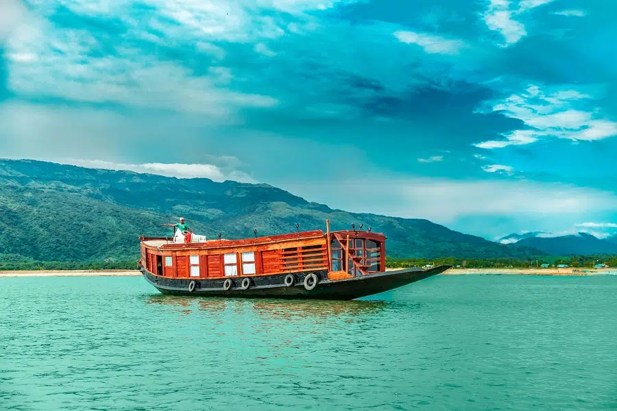 Traditional wooden boat with red accents floats on calm turquoise water under a partly cloudy sky; lush mountains and a sandy shore in the background.