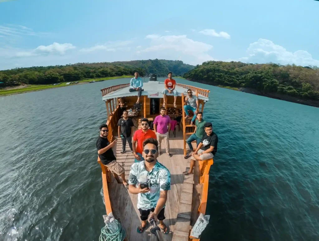 A group of friends smiling on a wooden boat in a scenic Rangamati Kaptai lake with lush greenery under a bright blue sky, conveying joy and camaraderie.