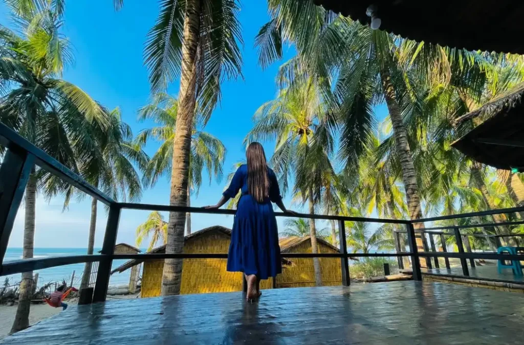 A woman in a blue dress stands barefoot on a wooden deck, gazing at palm trees and rustic huts under a clear blue sky, conveying serenity.