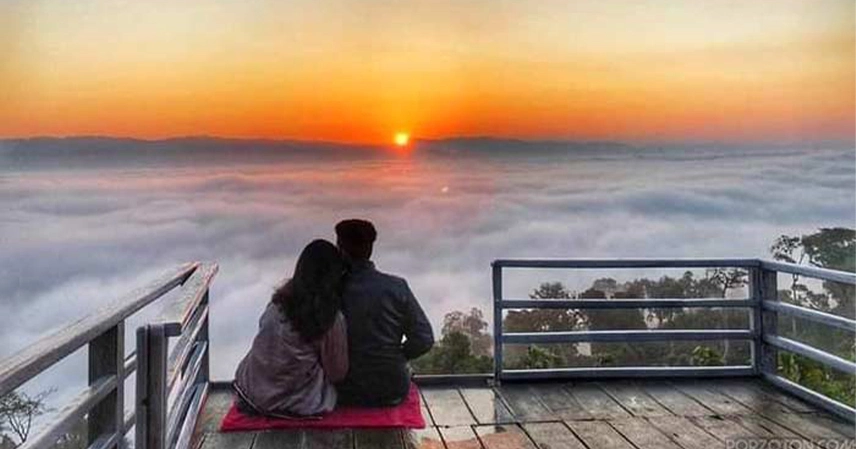 A couple sits on a red mat on a wooden balcony, watching a sunrise over a vast sea of clouds. The scene evokes tranquility and warmth.