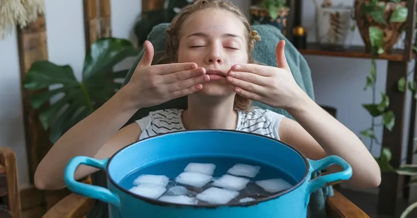 A person with closed eyes gently touches their face, seated indoors near a large blue pot filled with ice water. The setting is calm with plants around. বরফ ফেসিয়াল বা আইস ফেসিয়াল.