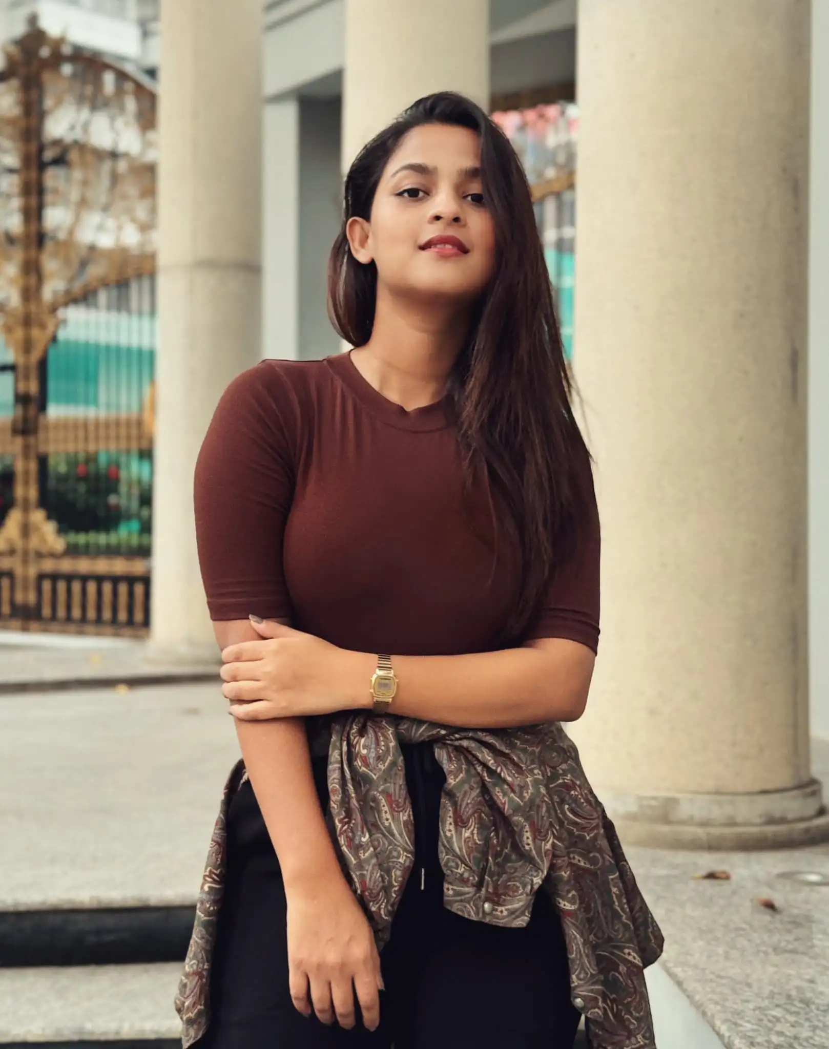 Young woman name Sadia Ayman in a brown shirt poses confidently outdoors. She has long hair and a watch on her wrist, with columns and ornate gates in the background.