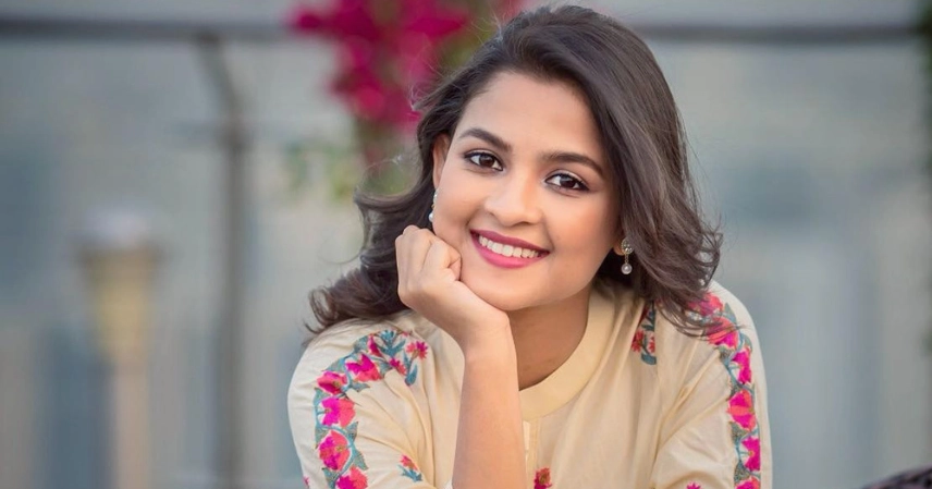 Smiling woman with dark hair and earrings, resting her chin on her hand. She's wearing a cream-colored top with vibrant floral patterns, exuding warmth. সাদিয়া আয়মানের ভাইরাল এআই ডিপ ফেক ভিডিও! সত্যটা জানুন