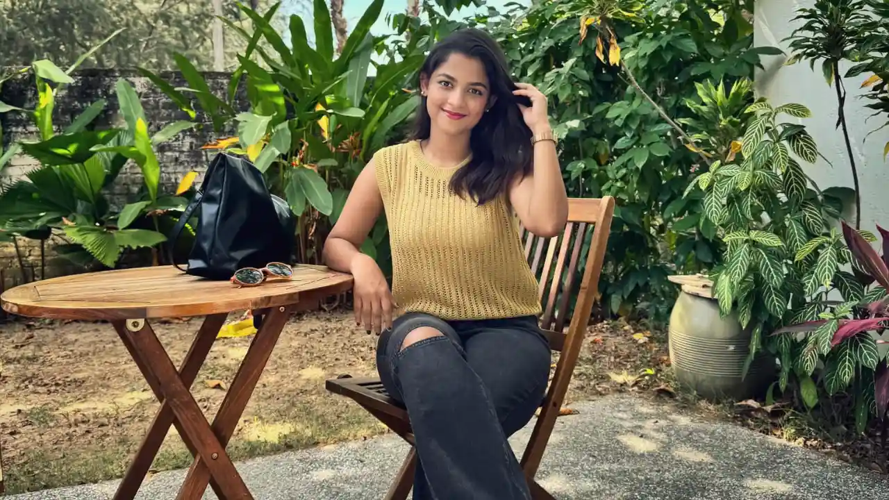 A woman sits at a wooden table in a lush garden, wearing a yellow top and black jeans. She smiles while touching her hair. A black bag and sunglasses are on the table.