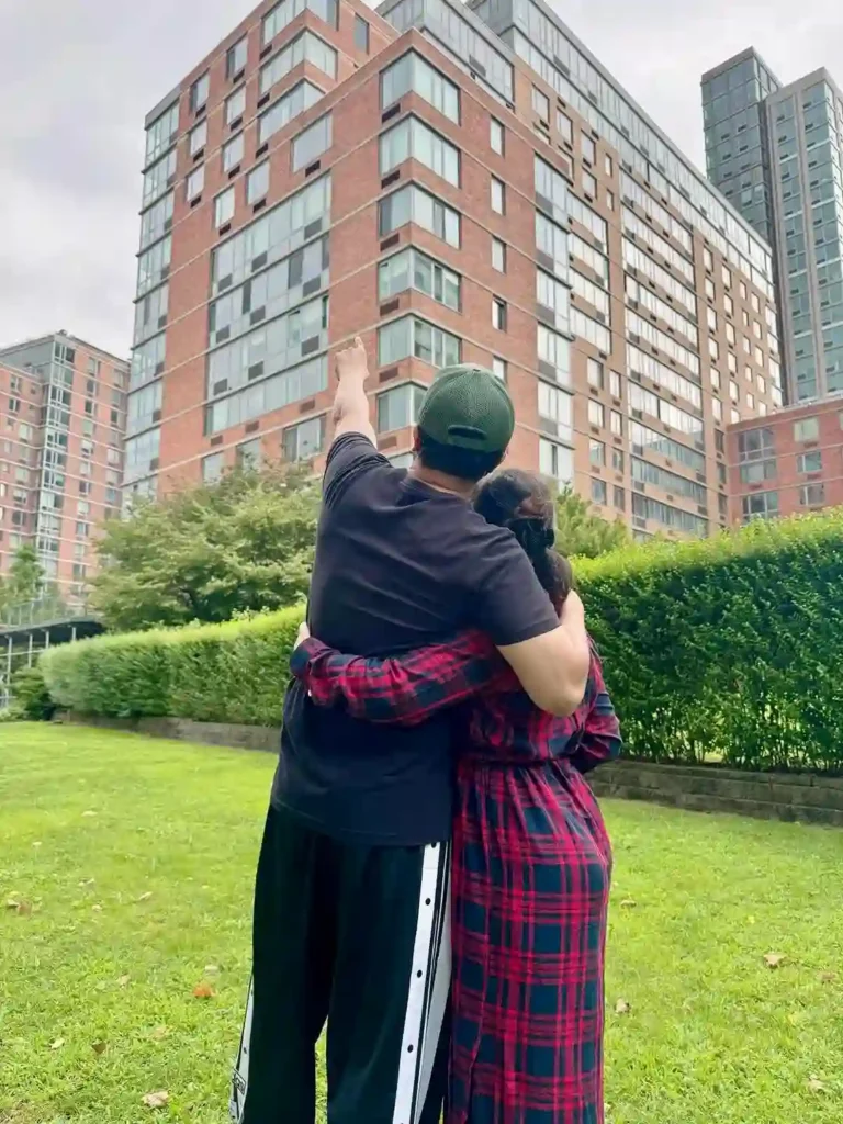 A couple stands on a grassy lawn, embracing and looking up at a tall brick building. One person is pointing towards the structure, evoking curiosity and excitement.