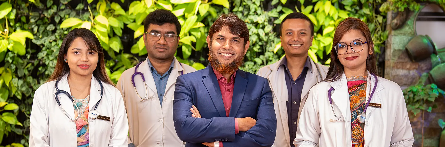 A group of five people stands confidently in front of a lush green wall. Four wear white lab coats with stethoscopes, and one wears a blue suit named Dr. Muhammad Jahangir Kabir, smiling.