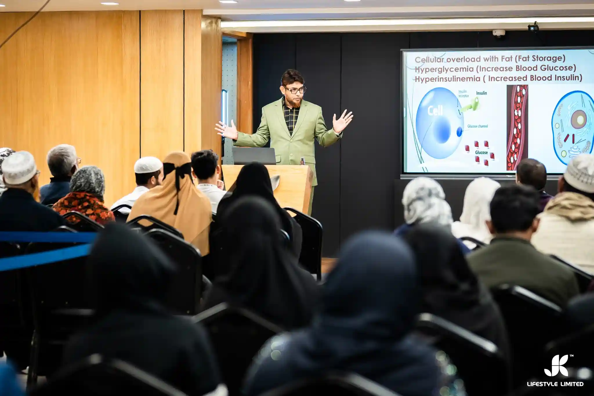 A man named Dr. Jahangir Kabir in a green suit gestures while addressing an audience in a lecture room. A presentation slide on cellular metabolism displays behind him. Attendees look attentive.