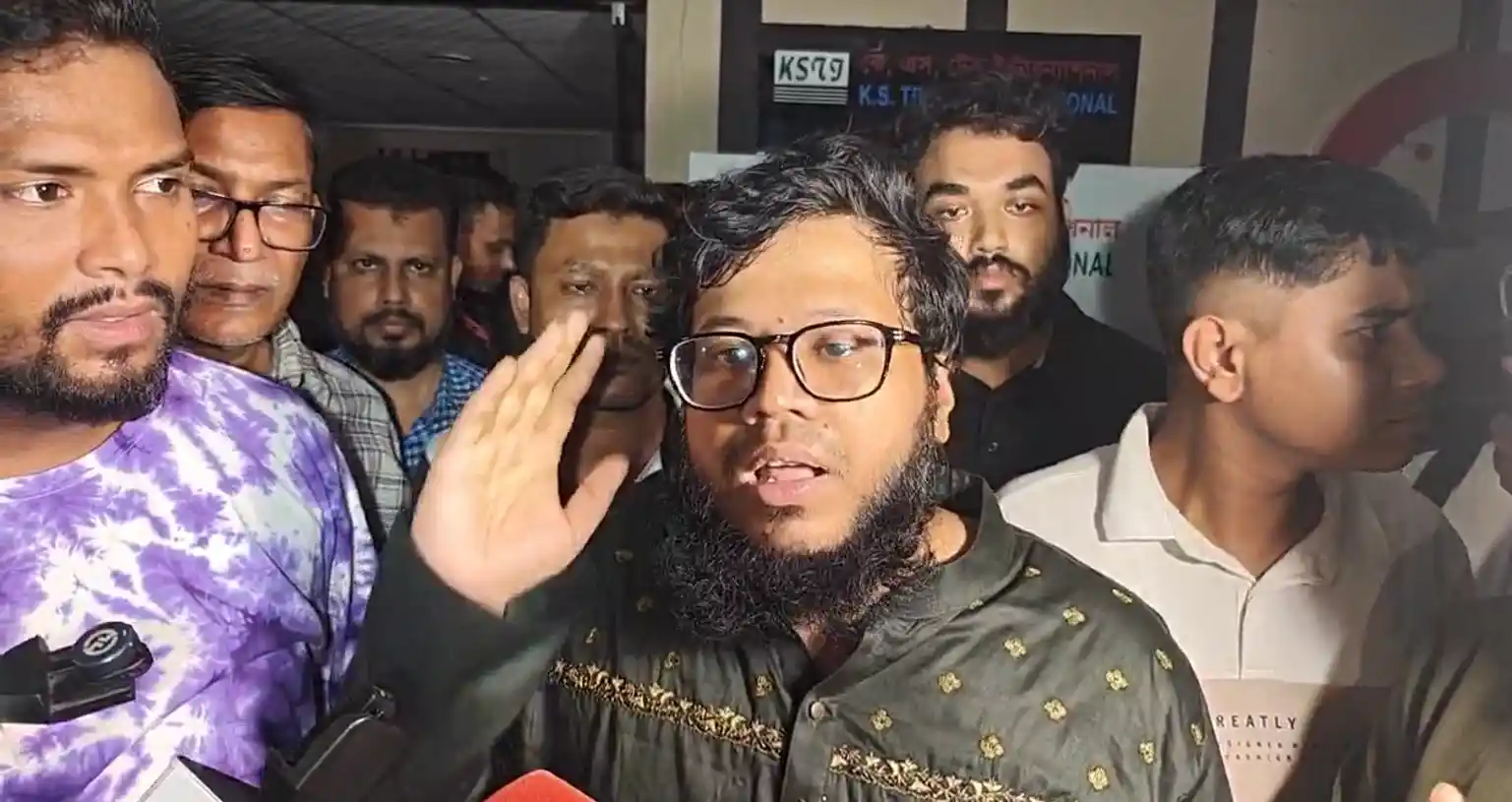 A group of men, one in a green embroidered shirt speaking passionately, surrounded by others in casual attire, appear focused and attentive. “ফ্লাইট এক্সপার্ট”