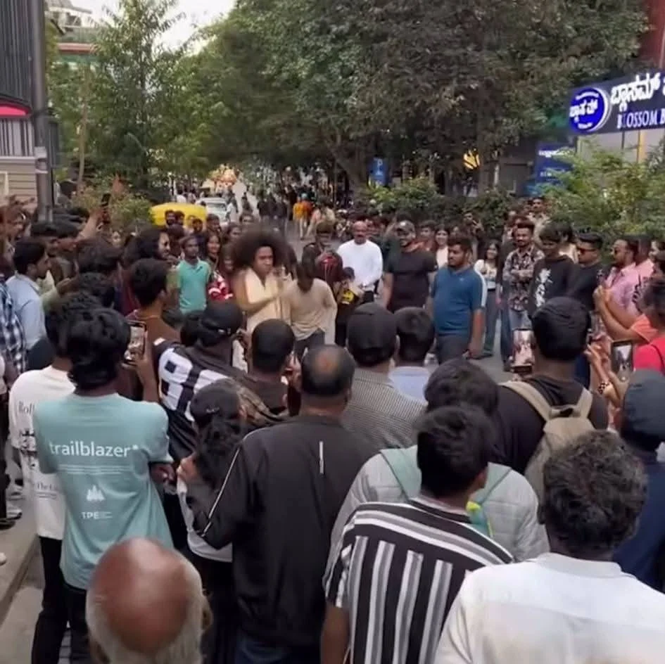 A lively street performance with dancers surrounded by a large, engaged crowd. Viewers hold up phones to capture the moment. The scene is vibrant and energetic.