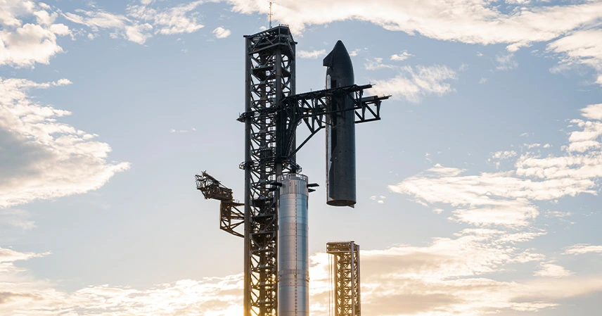 A large rocket stands vertically against a backdrop of a cloudy sky during sunset, casting a soft, glowing light. The scene conveys a sense of anticipation and innovation. স্পেসএক্স বিপর্যয়.