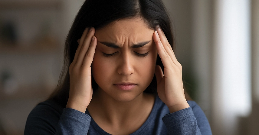 A woman with closed eyes holds her temples, showing discomfort. She wears a blue shirt, her expression indicates a headache or anxiety/stress. Blurred background.