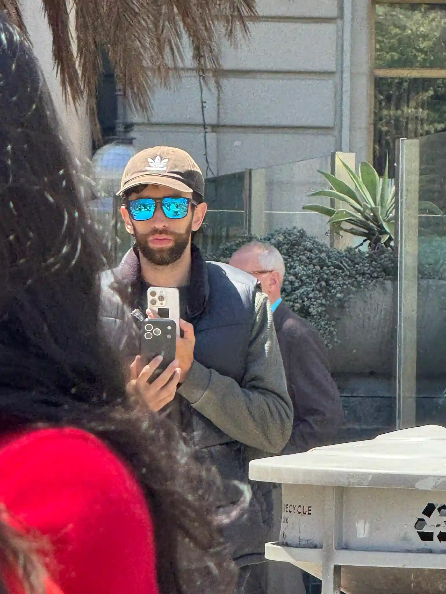 A man wearing a cap and reflective sunglasses takes a photo with his iphone 17 pro max phone. He stands in a sunny outdoor setting, with plants and a gray wall behind him.