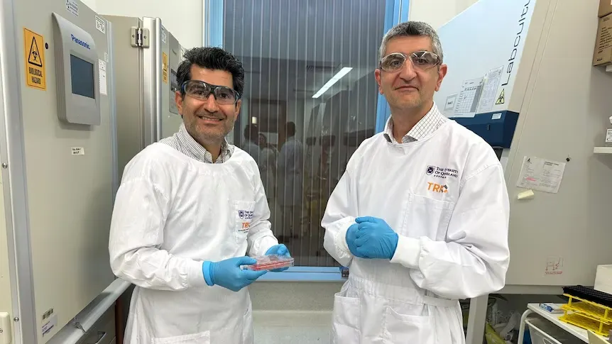 Two scientists in white lab coats and safety goggles stand in a lab, smiling. Both wear blue gloves and are positioned near lab equipment, conveying a sense of teamwork.
