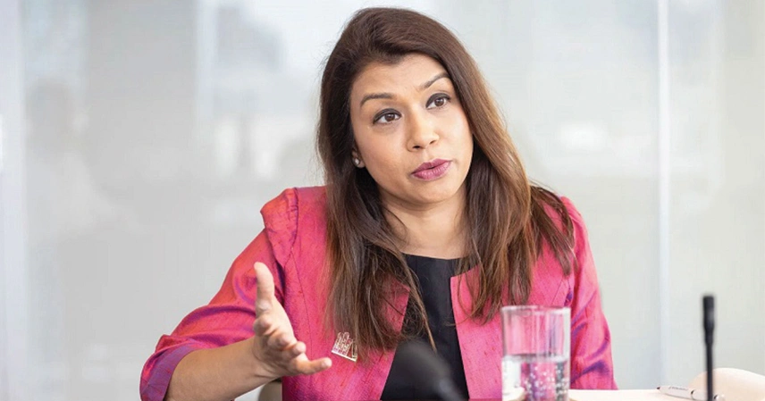 A person with long brown hair and wearing a pink blazer gestures with one hand, appearing thoughtful. A glass of water is on the table in front of them. টিউলিপ সিদ্দিক.