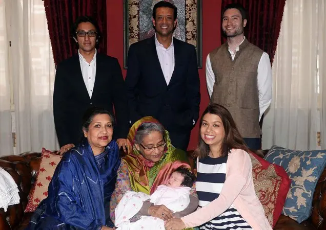 A group portrait showing three men standing at the back, dressed formally. Three women are seated in front, one holding a baby, all smiling warmly. The setting is cozy, with patterned curtains and cushions, radiating a familial, joyful atmosphere.