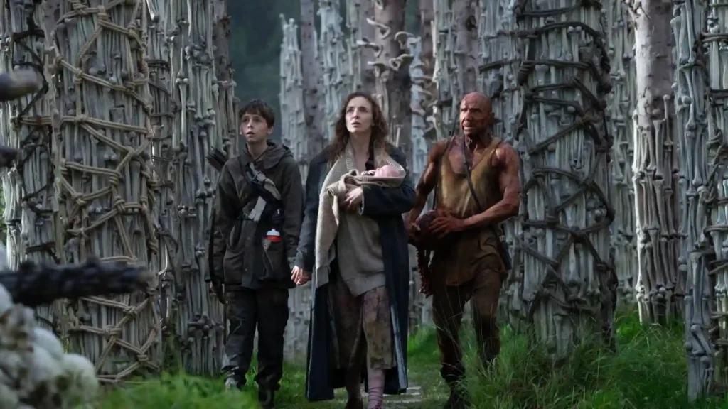 A group walks through a forest of tall, alien-like structures. A woman cradles a baby, flanked by a young boy and a muscular man. The scene conveys tension.