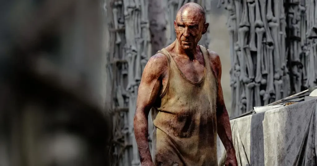 A rugged, bald man in a worn tank top stands against a textured, industrial background. His intense expression and dirt-covered skin convey tension. 28 years later the bone temple trailer.