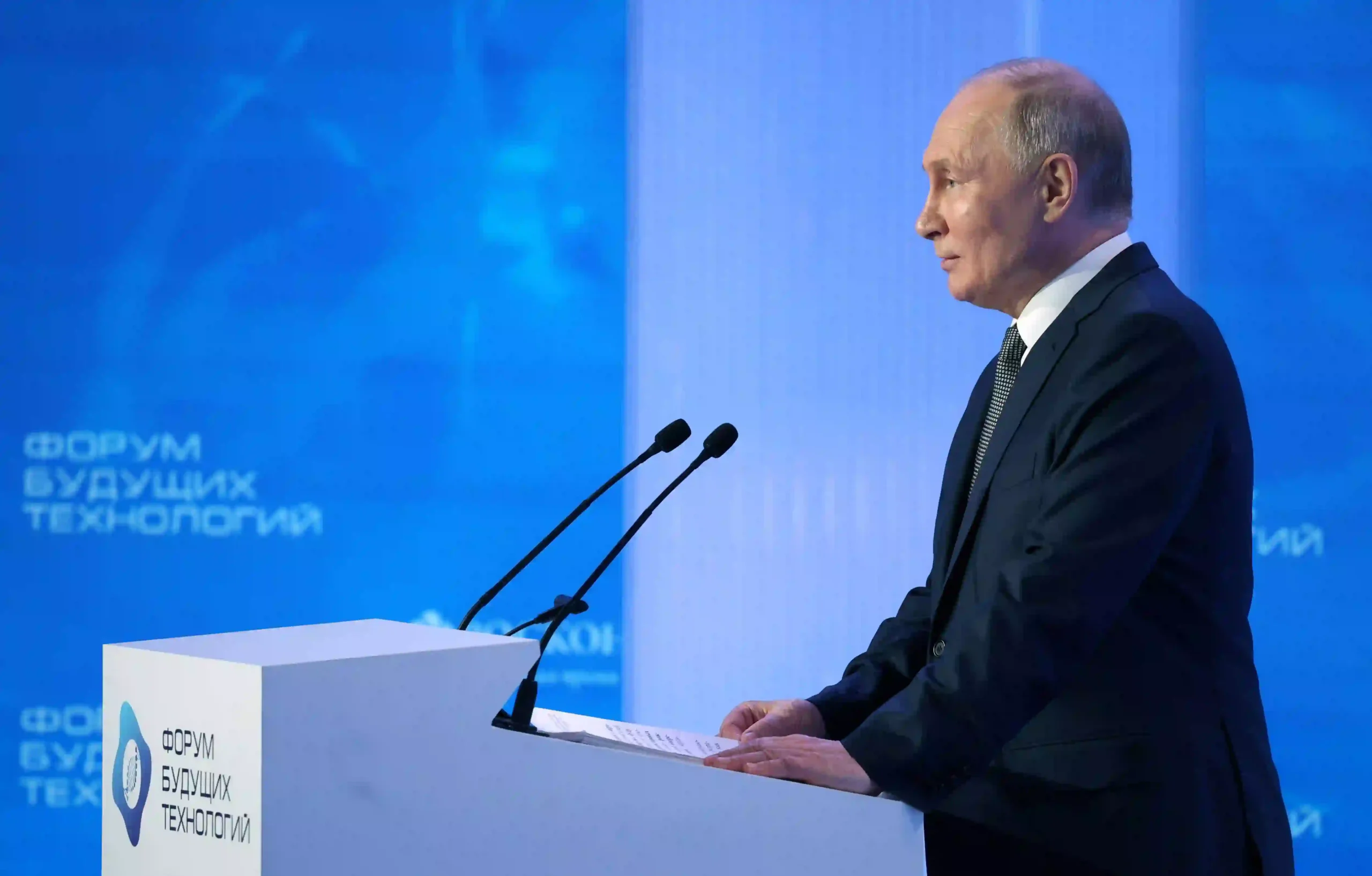 A speaker stands at a podium with microphones, addressing an audience at a technology forum, set against a blue backdrop.
